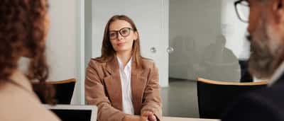 A young woman in a corduroy blazer sits across from two people during a job interview.
