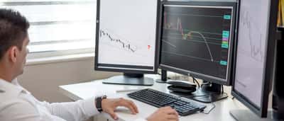 A financial professional analyzes market charts on multiple computer monitors in an office.