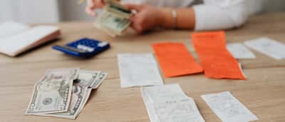 A person counts cash on a wooden desk scattered with shopping receipts and a blue calculator.