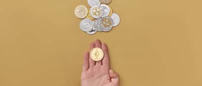 A hand holding a physical cryptocurrency coin above a scattered pile of digital currency coins on a bright yellow background.