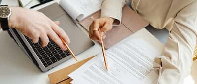 Overhead view of two people using pens to point out specific details on a printed document next to a laptop.