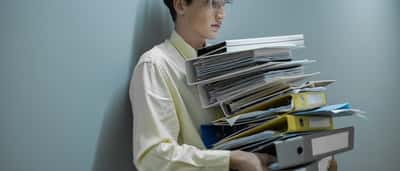 An office worker carries a massive, heavy stack of colourful document folders in a modern workplace.