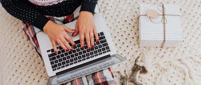 A person wearing a festive sweater and plaid pants types on a laptop next to a wrapped holiday gift on a knitted blanket.
