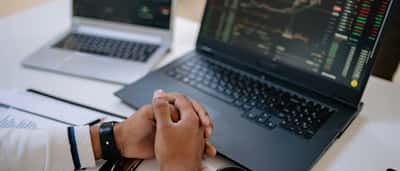 A person sits at a desk with folded hands, monitoring financial charts on two laptop screens.
