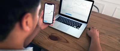 A person sits at a wooden desk analyzing financial market charts simultaneously on a smartphone and a laptop.