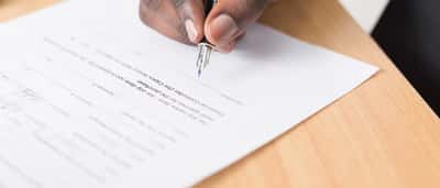 A close-up of a person using a fountain pen to sign a financial document on a wooden desk.