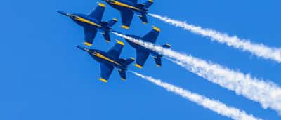 Four U.S. Navy Blue Angels F/A-18 fighter jets soar in tight formation with white smoke trails against a bright blue sky.