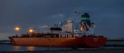 A large oil tanker ship is illuminated against a dark, moody sky at an industrial port.