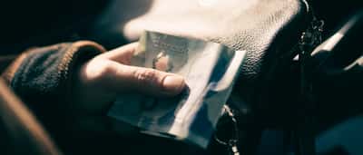 A person's hand holding a blue Canadian five-dollar bill next to a leather handbag.