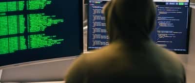 An anonymous person in a hoodie sits in a dark room working on computer monitors displaying lines of code.