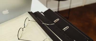 A pair of wire-rimmed reading glasses resting on a document over a black binder next to a silver laptop.