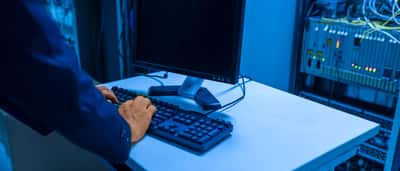 A person's hands type on a computer keyboard at a workstation inside a blue-lit server room.