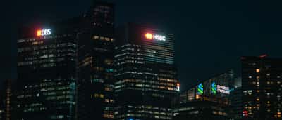 Illuminated logos of major banks shine from skyscrapers against a dark night sky.