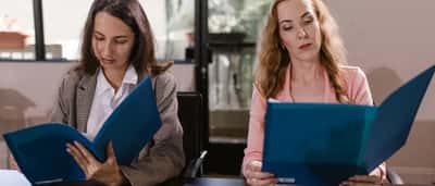 Two professional women sit side-by-side at a conference table, each reading from identical blue folders.