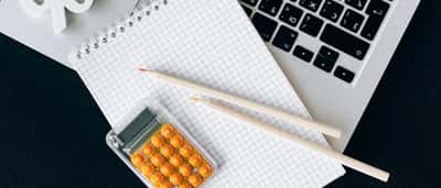 A top-down view of a modern desk featuring a laptop, grid notebook, orange calculator, and a white percent symbol.