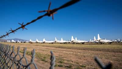 A row of parked military aircraft sitting idle in a desert boneyard, viewed through a barbed wire fence.