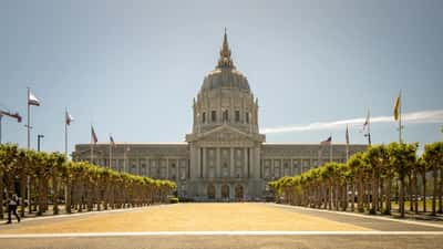The dome and facade of San Francisco City Hall stand at the end of a long, tree-lined plaza.