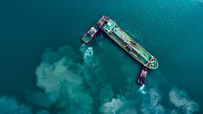 A top-down aerial view of an oil tanker being guided by two tugboats in deep turquoise waters.