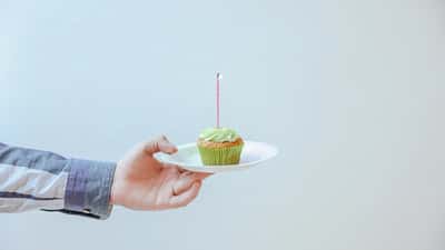 A hand in a striped button-down shirt holds a small plate with a green cupcake and a single lit candle.