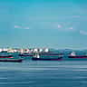 Several oil tankers wait on the water in front of a coastal fuel storage terminal with large white tanks.