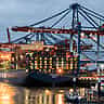 A massive cargo ship loaded with shipping containers is docked at an illuminated port terminal at dusk.