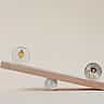A silver Ethereum coin and a silver Bitcoin coin balancing on opposite ends of a wooden seesaw against a beige background.