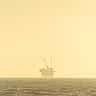 An offshore oil platform sits on the horizon under a golden, hazy sky.