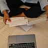 A person's hands hold printed checks over a desk with a laptop.