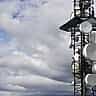 A tall telecommunications tower equipped with large white satellite dishes stands against a cloudy sky.