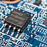 A black microchip is soldered onto a blue printed circuit board among other small electronic components.
