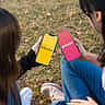 Two people sit outdoors holding smartphones that display the Bumble and Tinder dating apps.