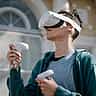 A young person uses a white Meta Quest virtual reality headset and hand controllers outdoors in the sunlight.