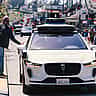 A white Waymo self-driving vehicle with roof-mounted sensors navigates a city street.