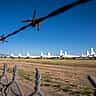 A row of parked military aircraft sitting idle in a desert boneyard, viewed through a barbed wire fence.
