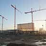 Multiple towering cranes stand over a massive building framework at a large-scale construction site at dusk.