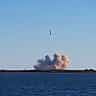 A rocket launches into a clear blue sky, leaving a thick trail of white smoke.