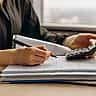 A person uses a pen and a calculator to review a stack of financial documents.