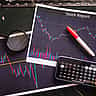 A printed stock report showing candlestick charts, resting on a dark surface alongside a magnifying glass, marker, and a calculator on a smartphone.