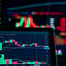 A digital trading chart is displayed on a screen in sharp focus, with a larger, brightly lit chart blurred in the background.