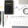 A flat lay of a desk featuring financial line charts, a dotted world map graphic, a calculator, and a magnifying glass.