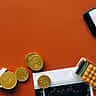A flat lay composition featuring a white percentage sign, gold coins, a calculator, and a smartphone displaying a financial chart on a warm orange background.