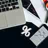 A white percentage symbol rests on a dark desk surrounded by a laptop, a smartphone displaying market data, and printed financial charts.