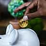 A hand places a gold physical Bitcoin token into the slot of a white ceramic piggy bank.