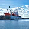 A large orange ship sits in a floating dry dock at an industrial shipyard under a bright blue sky.