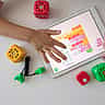 A child's hands interact with a block-based coding application on a digital tablet, surrounded by colorful cube toys.