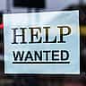 A red and white Help Wanted sign taped to the inside of a glass storefront window.
