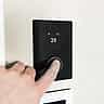 A close-up of a person's finger pressing the dial on a sleek, black digital smart thermostat mounted on a wall.