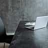 An empty black leather office chair sits next to a dark desk with an open laptop against a concrete wall.