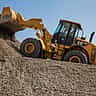 A yellow Caterpillar front loader carrying gravel up a steep mound under a clear blue sky.