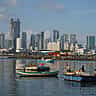 Traditional fishing boats float in a harbor with modern skyscrapers under construction in the background.